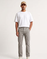 A man modeling a white crewneck t-shirt and light gray chino pants, paired with a beige baseball cap and white sneakers, against a plain off-white studio background.