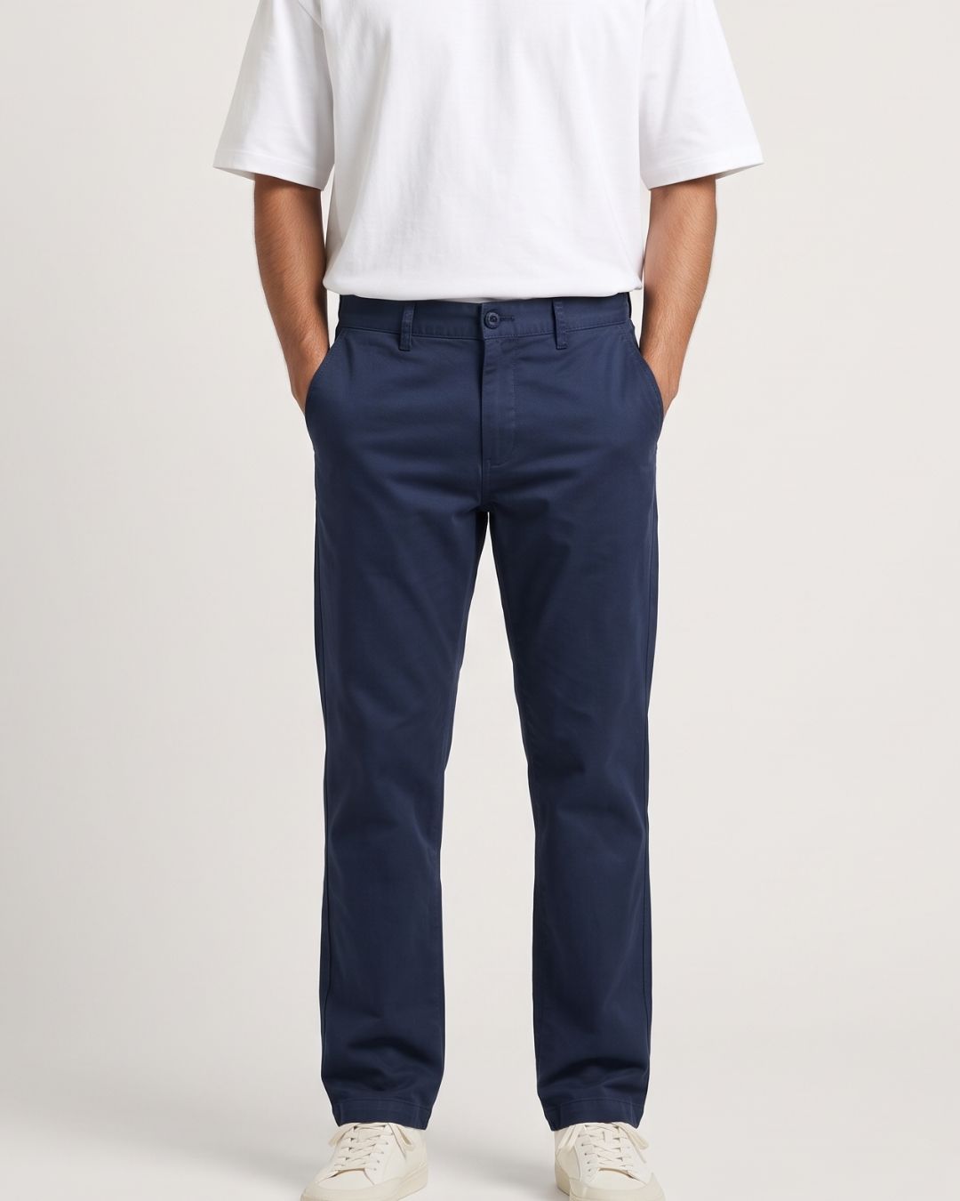 A person wearing navy blue chino pants, a white t-shirt, and white sneakers, standing against a light gray studio background.