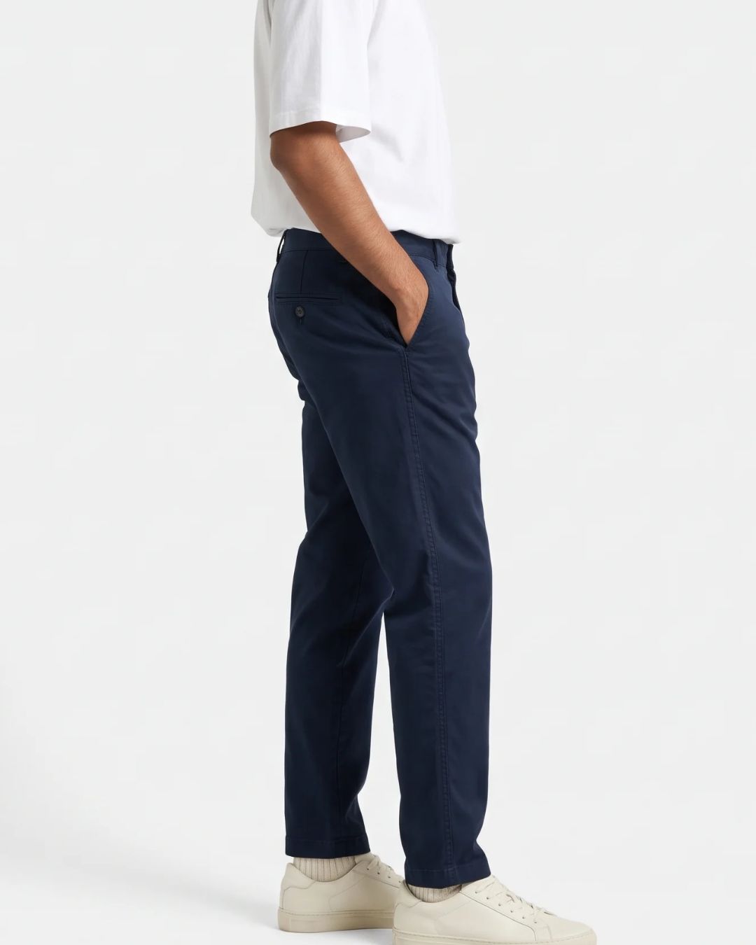 A side profile view of a person wearing navy blue trousers, a white t-shirt, and off-white sneakers against a plain white studio background.