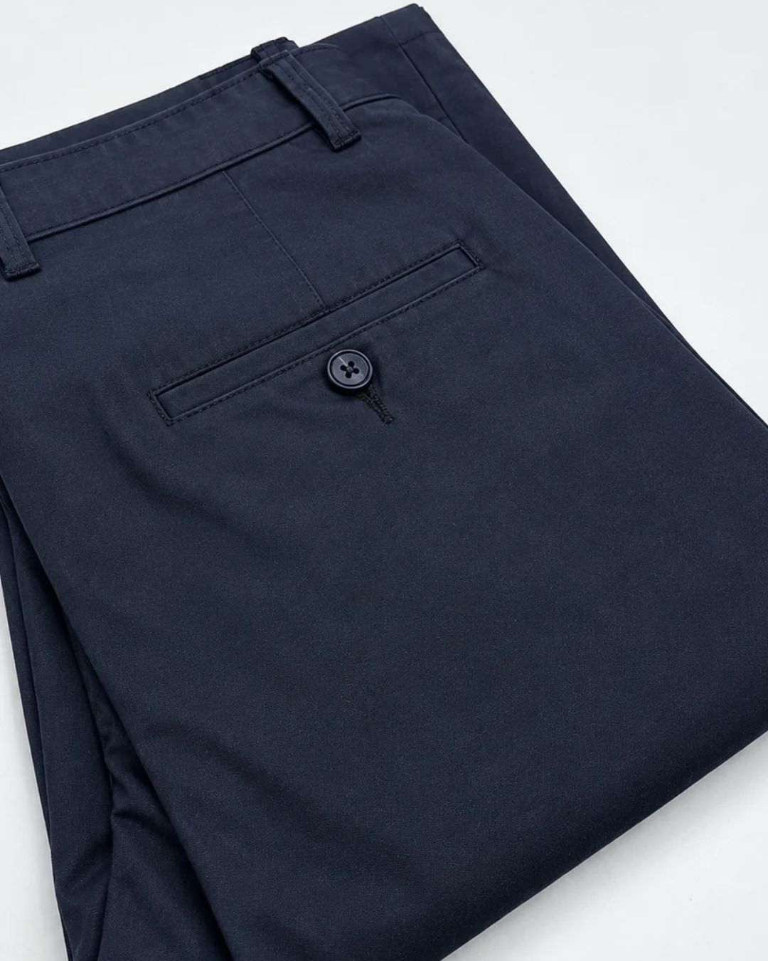 A close-up of folded navy blue trousers showing the waistband, belt loops, and a rear welt pocket with a button closure, photographed on a clean white background.