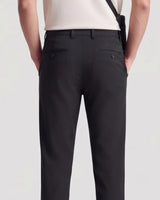 Rear view of a person wearing black tailored trousers with a cream top and a black shoulder bag strap visible against a light gray studio background.