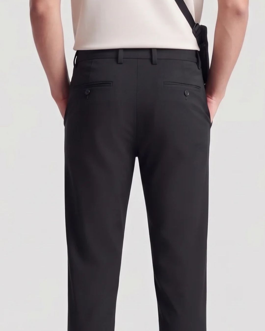 Rear view of a person wearing black tailored trousers with a cream top and a black shoulder bag strap visible against a light gray studio background.