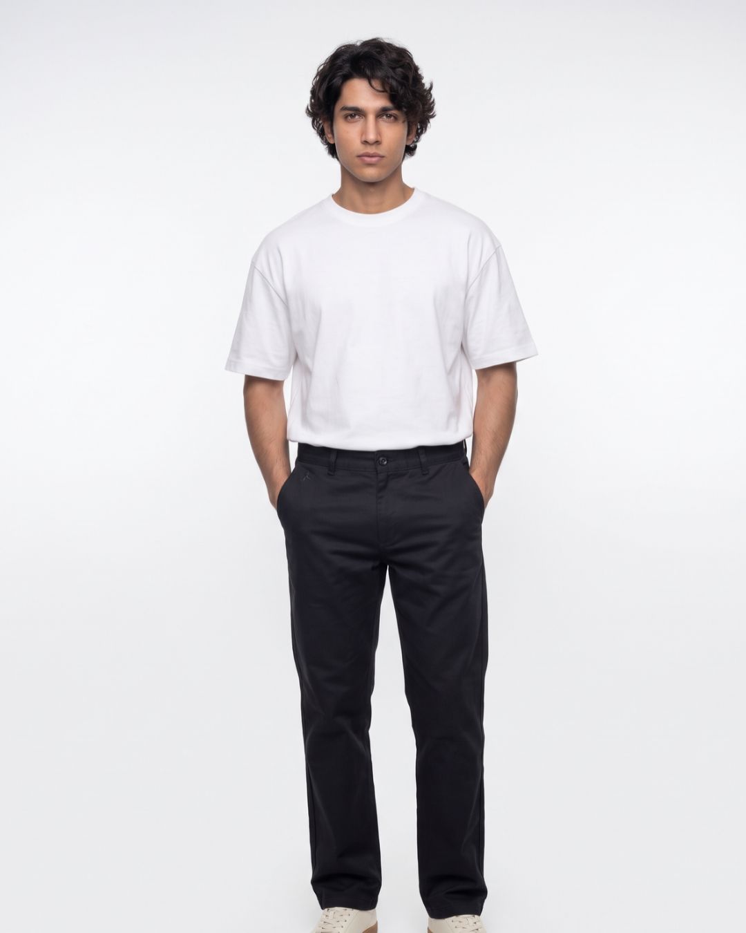 A man modeling a white crew-neck t-shirt and black trousers against a plain white studio background.
