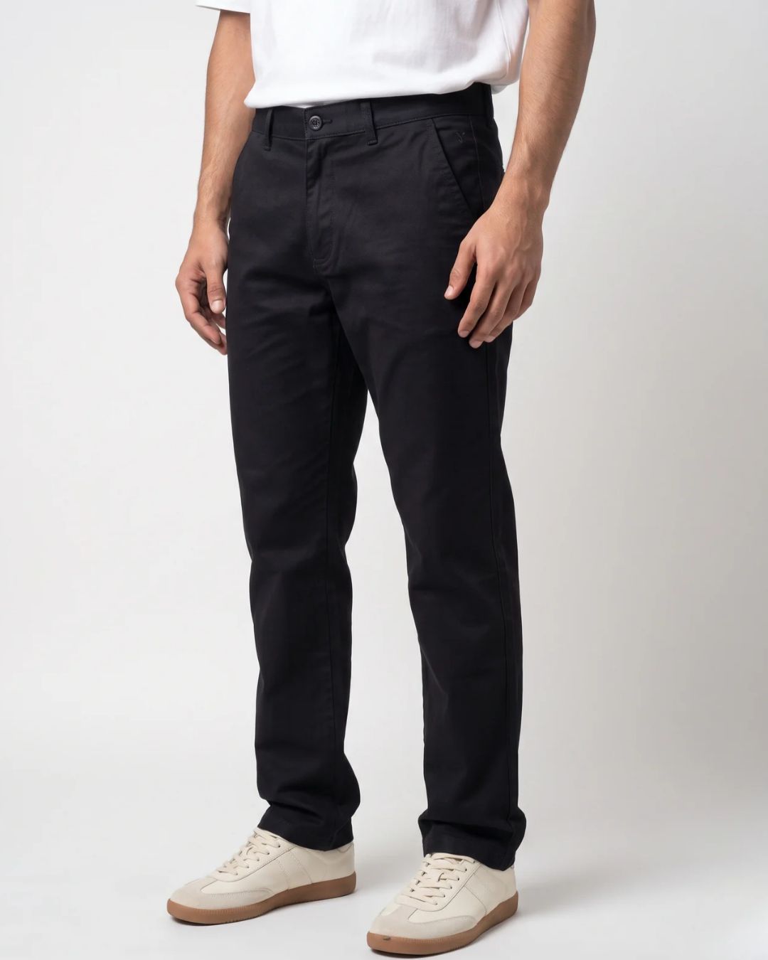 A person wearing slim black trousers, a white t-shirt, and beige sneakers, standing against a light gray studio background.