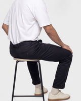 A man wearing black trousers, a white t-shirt, and off-white sneakers sits on a black metal stool against a light gray background.