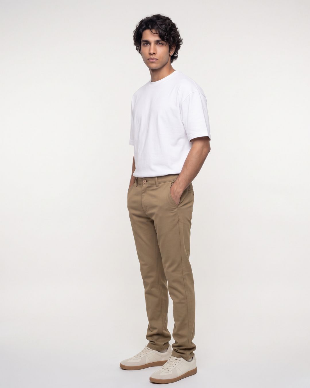 A man modeling a white crew-neck t-shirt, khaki brown slim-fit trousers, and off-white sneakers against a plain white studio background.