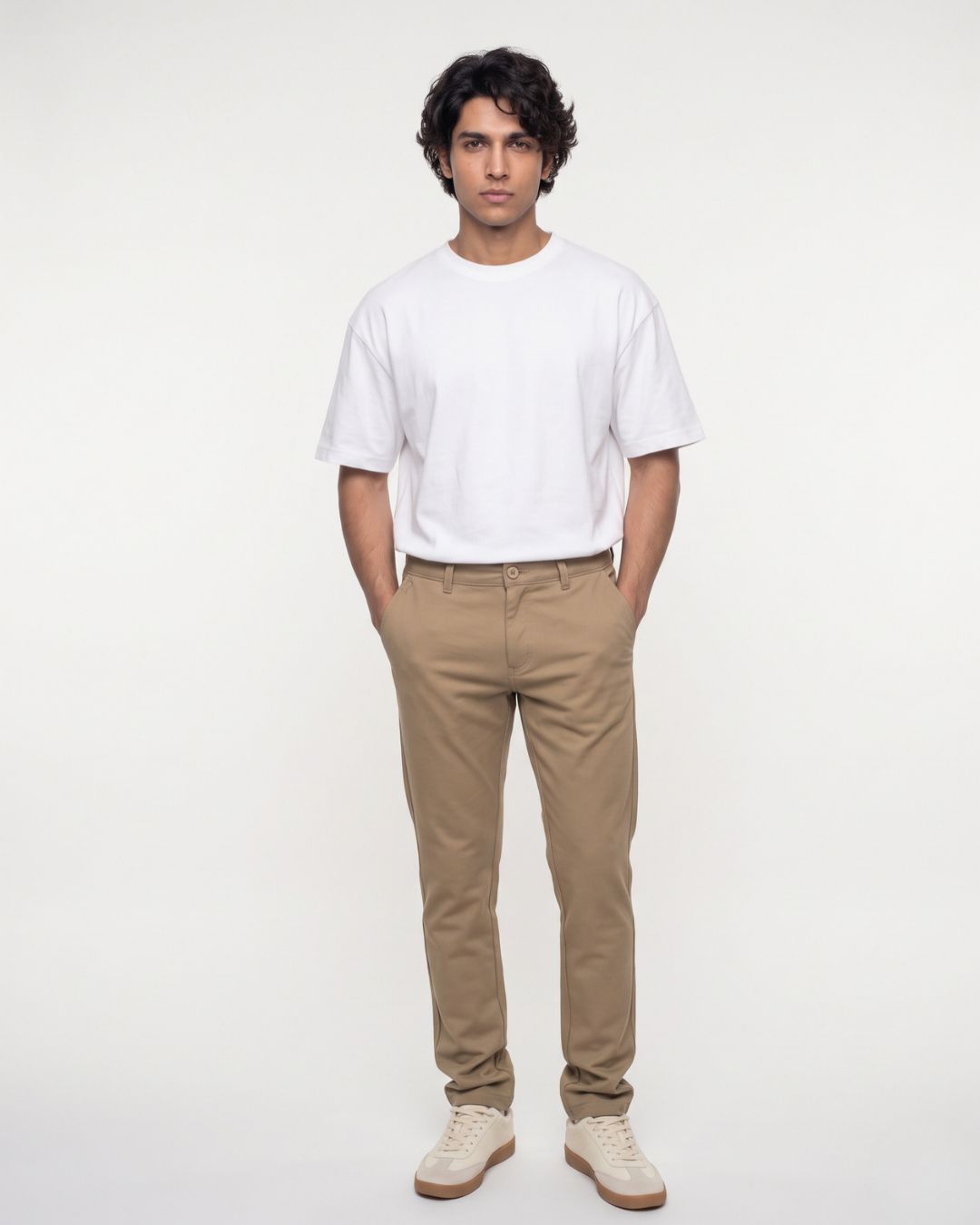 A person seated on a wooden stool wearing slim tan chinos, a white short-sleeve T-shirt, and off-white sneakers against a plain light background