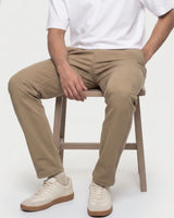 A person seated on a wooden stool wearing slim tan chinos, a white short-sleeve T-shirt, and off-white sneakers against a plain light background.