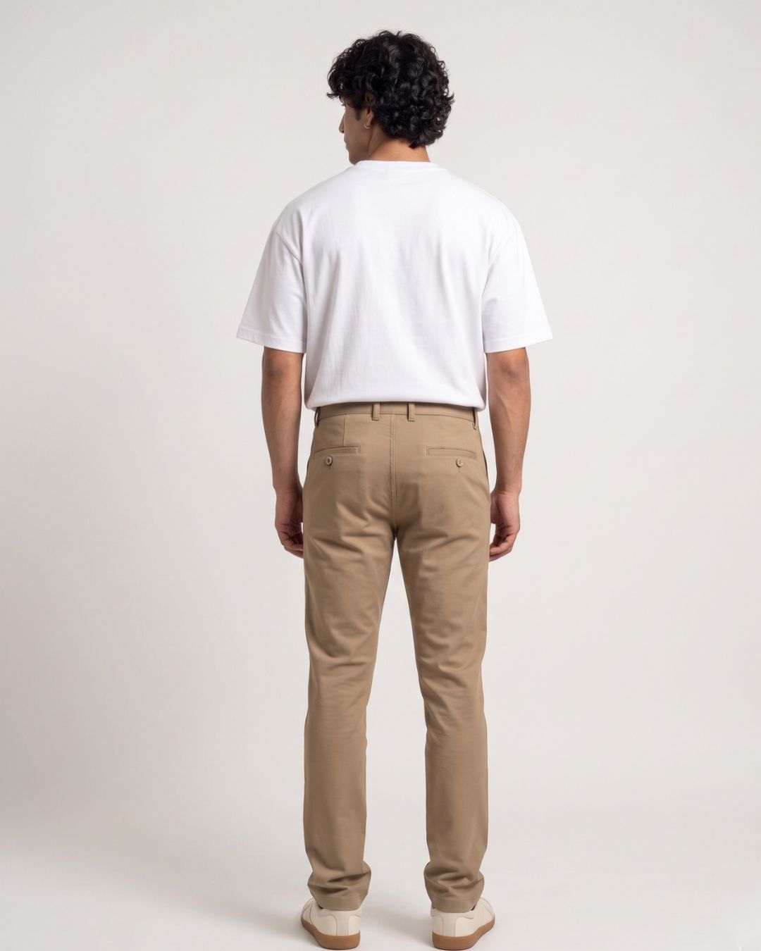 A person seated on a wooden stool wearing slim tan chinos, a white short-sleeve T-shirt, and off-white sneakers against a plain light background