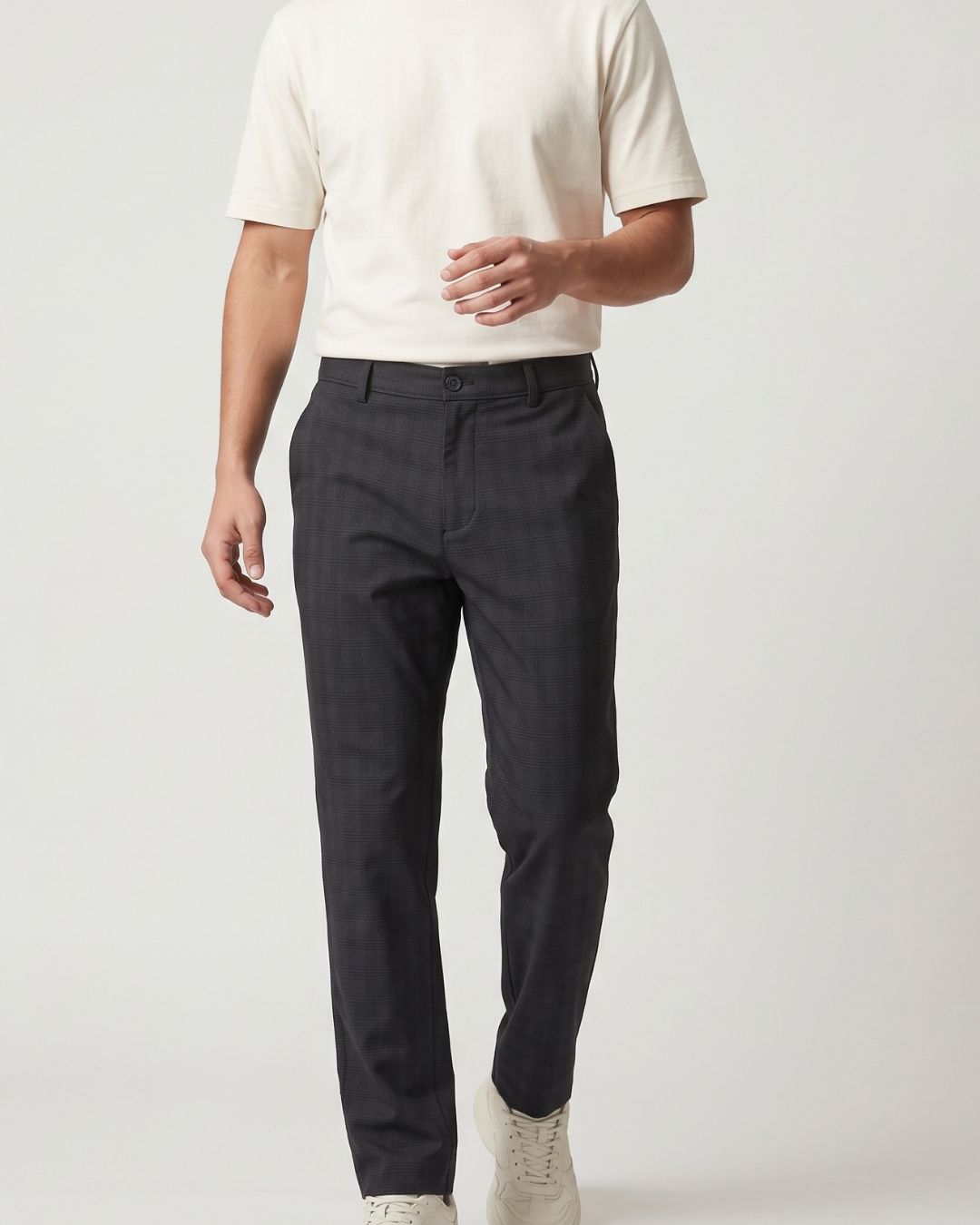 A man wearing dark charcoal gray slim-fit trousers, a cream short-sleeve t-shirt, and off-white sneakers, shown from the waist down against a light gray studio background.