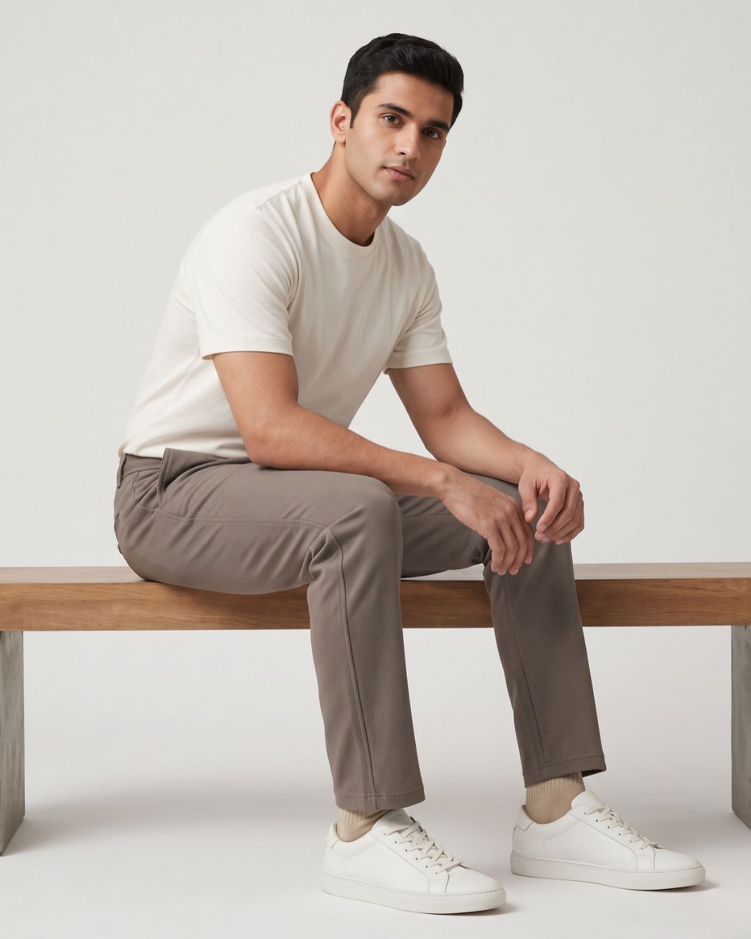 A man sitting on a wooden bench wearing a light beige crew-neck t-shirt, taupe trousers, and white sneakers against a plain light gray studio background.