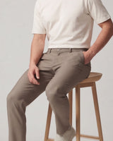 A man wearing slim-fit taupe trousers and a cream t-shirt sits on a wooden stool against a light gray background.