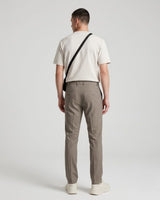 A man viewed from behind wearing a cream t-shirt, taupe trousers, and white sneakers, with a black crossbody bag slung over his shoulder, set against a plain light gray studio background.