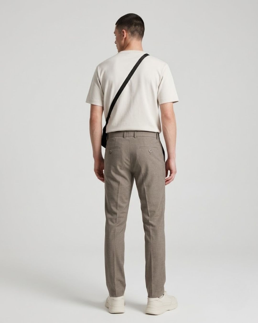 A man viewed from behind wearing a cream t-shirt, taupe trousers, and white sneakers, with a black crossbody bag slung over his shoulder, set against a plain light gray studio background.