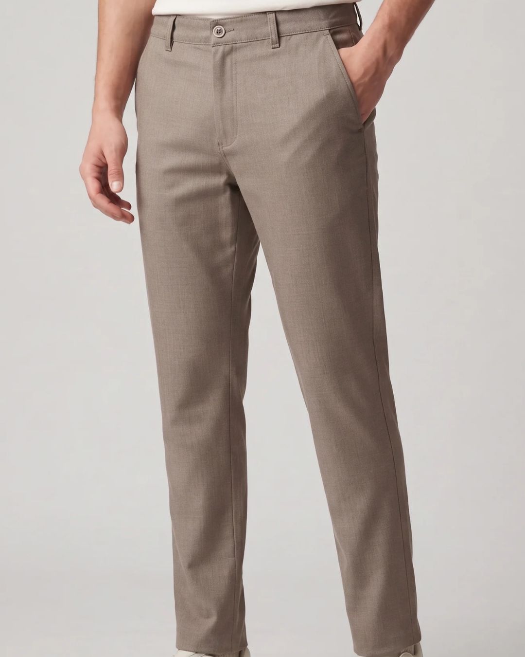 A person wearing slim-fit taupe trousers, shown from the waist down against a light gray studio background.