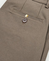 Close-up of a pair of taupe-brown trousers showing the back pocket with a single button closure and a small gold accent tab.