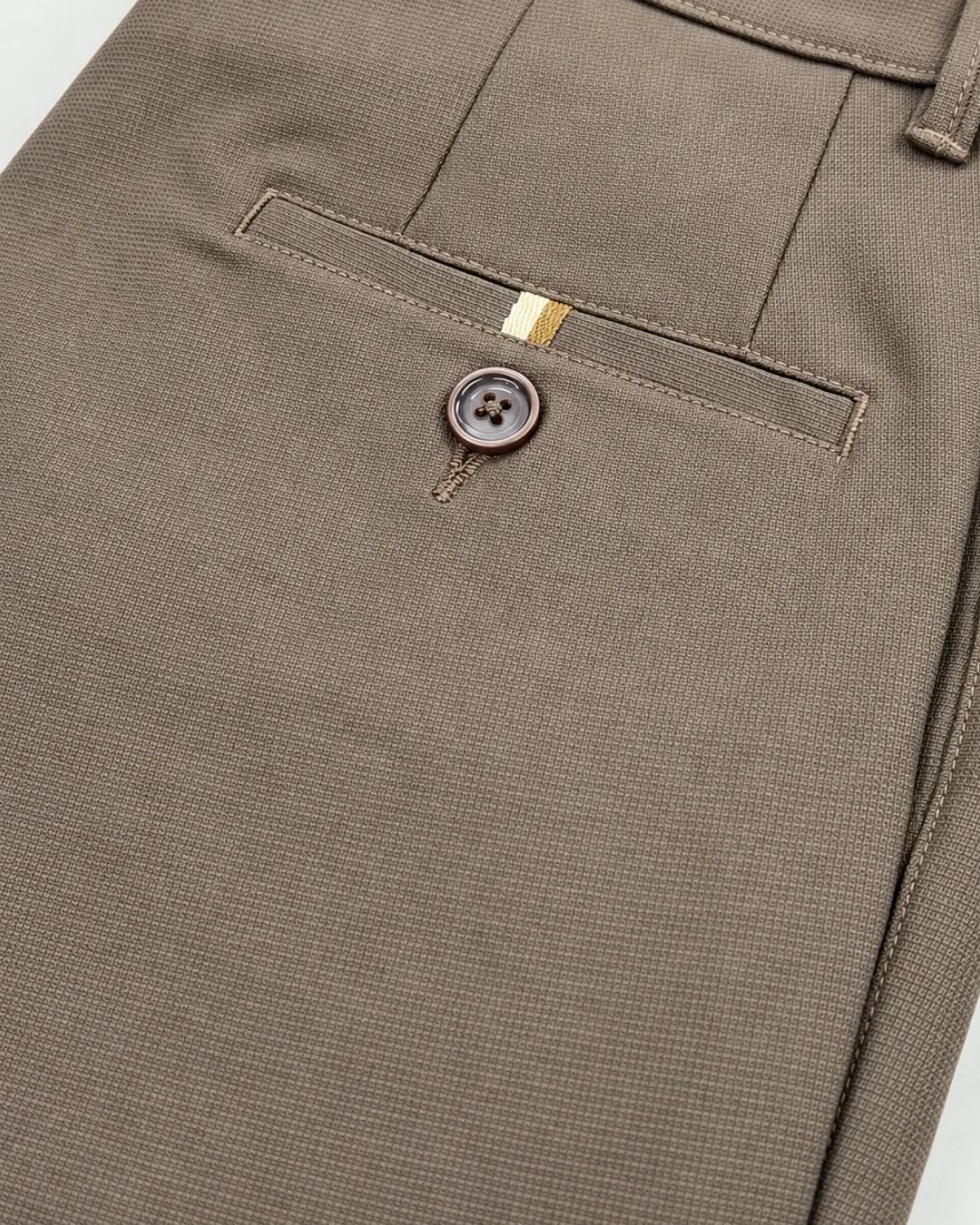 Close-up of a pair of taupe-brown trousers showing the back pocket with a single button closure and a small gold accent tab.