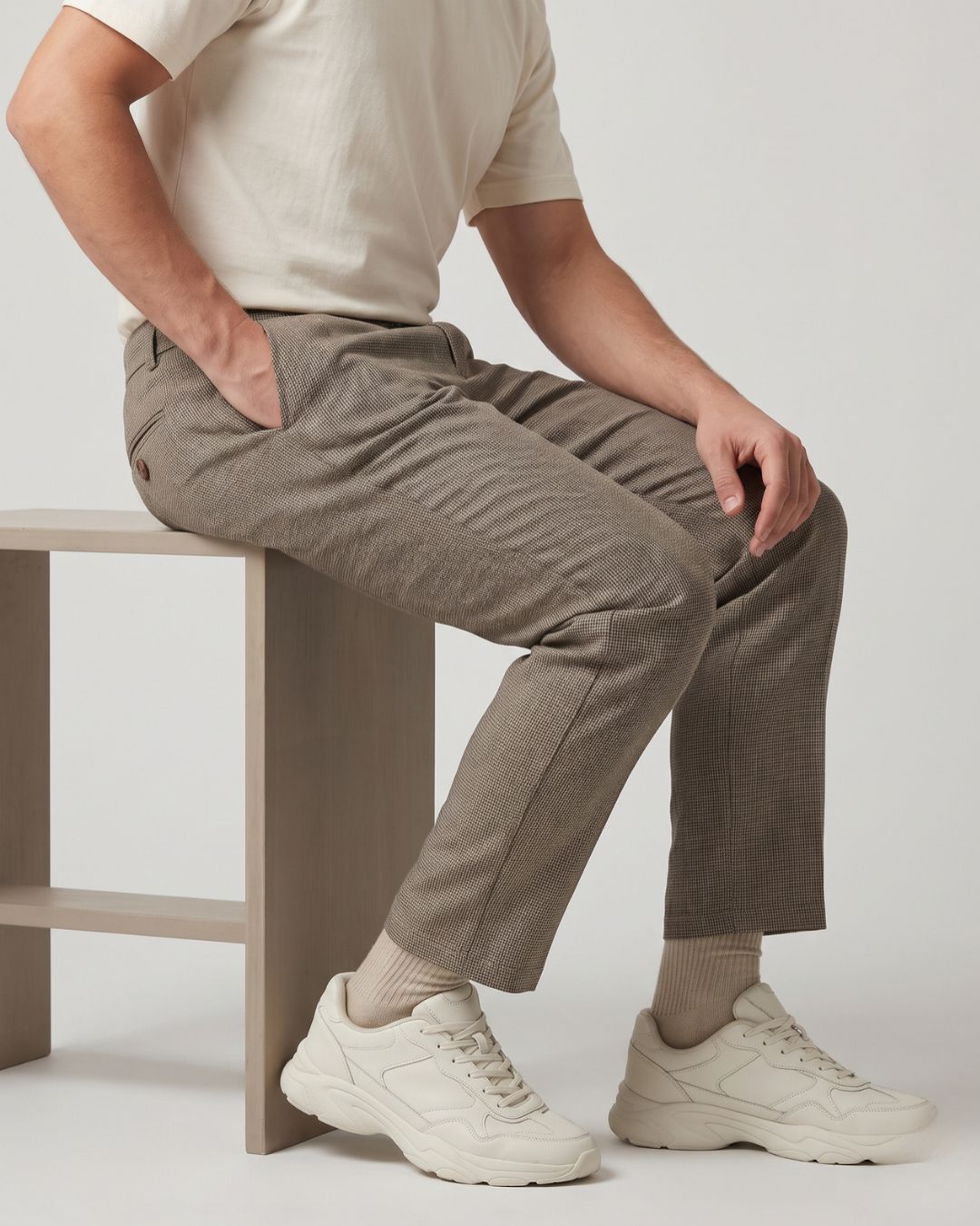 A model wearing textured taupe-brown trousers, a cream t-shirt, and chunky off-white sneakers sits on a minimalist wooden stool against a light gray studio background.