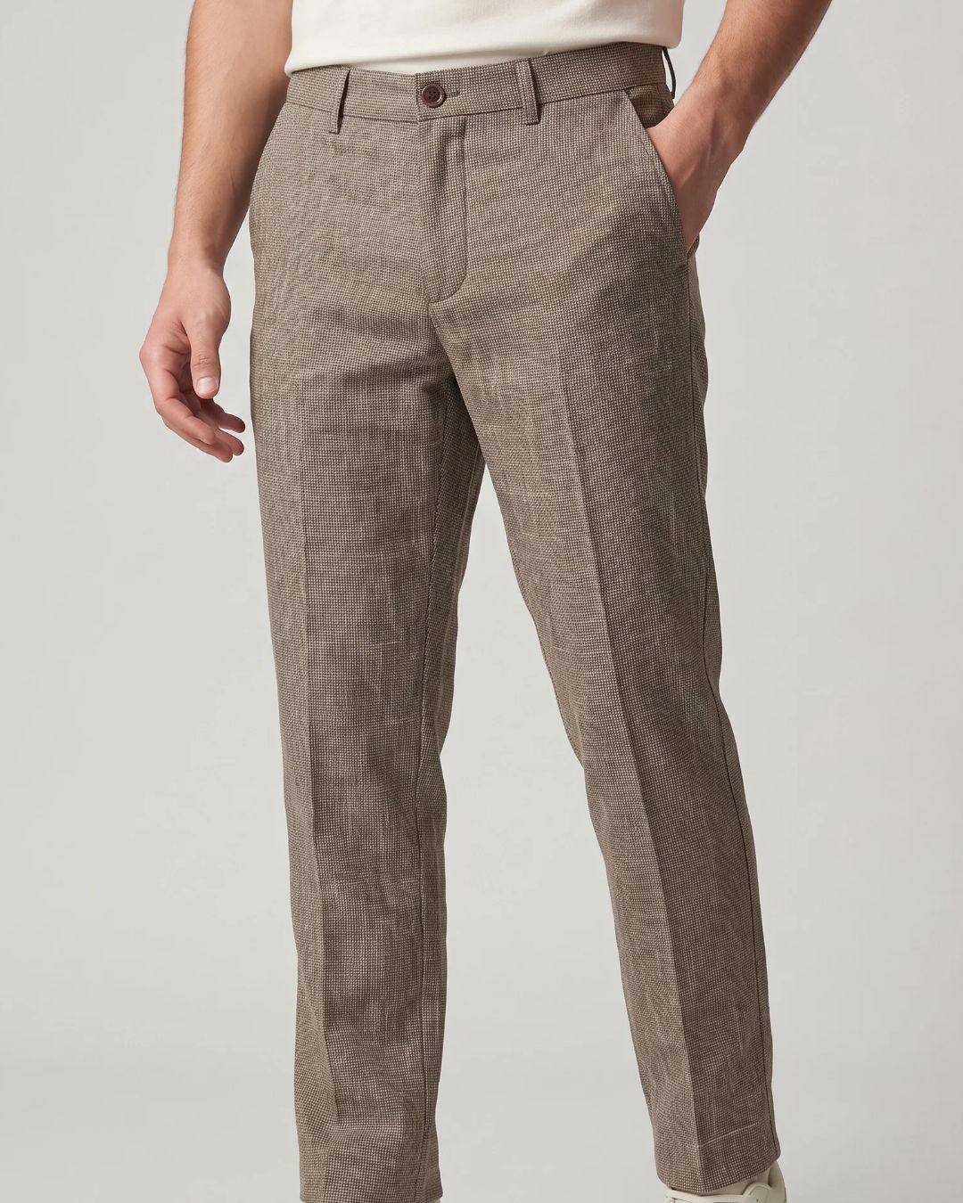 A person wearing slim-fit trousers in a taupe-brown houndstooth pattern, paired with a white top, shown from the waist down against a light gray studio background.