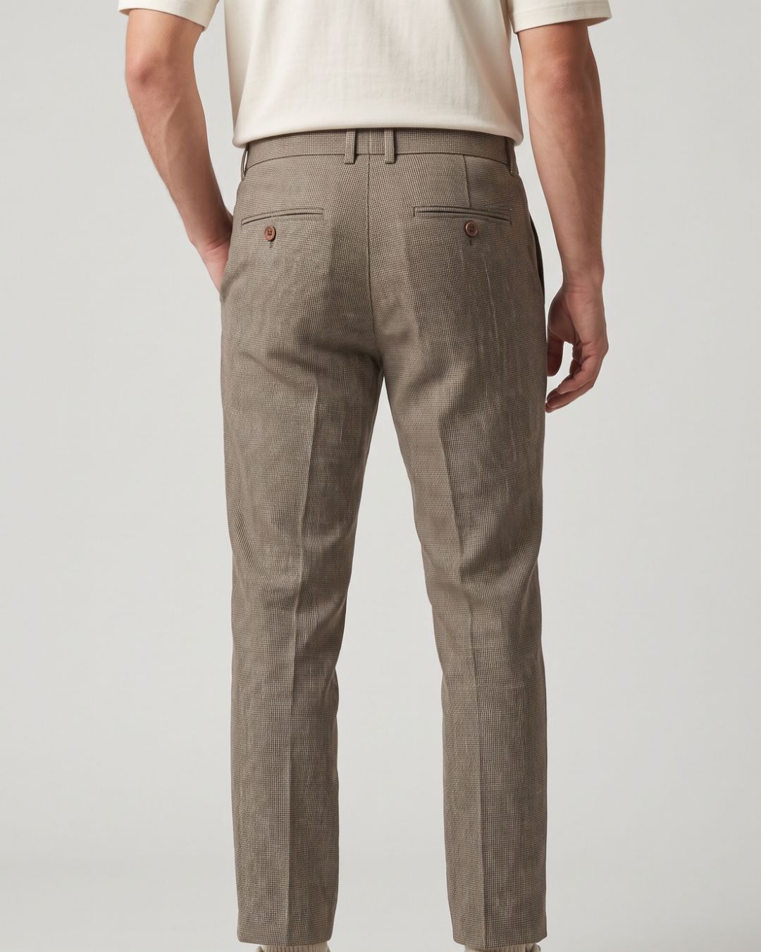 Rear view of a person wearing slim-fit trousers in a warm taupe-brown color, paired with a cream short-sleeve top, against a light gray studio background.