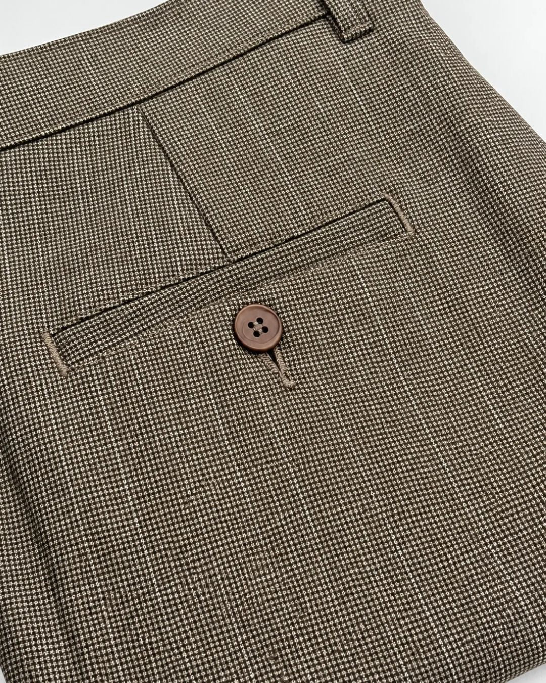 Close-up of a brown and beige micro-check patterned fabric, showing a buttoned welt pocket and belt loop detail.