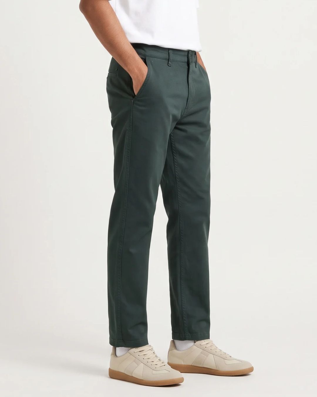 A person wearing dark green slim-fit chinos with a white t-shirt and beige sneakers, shown from the waist down against a light gray studio background.