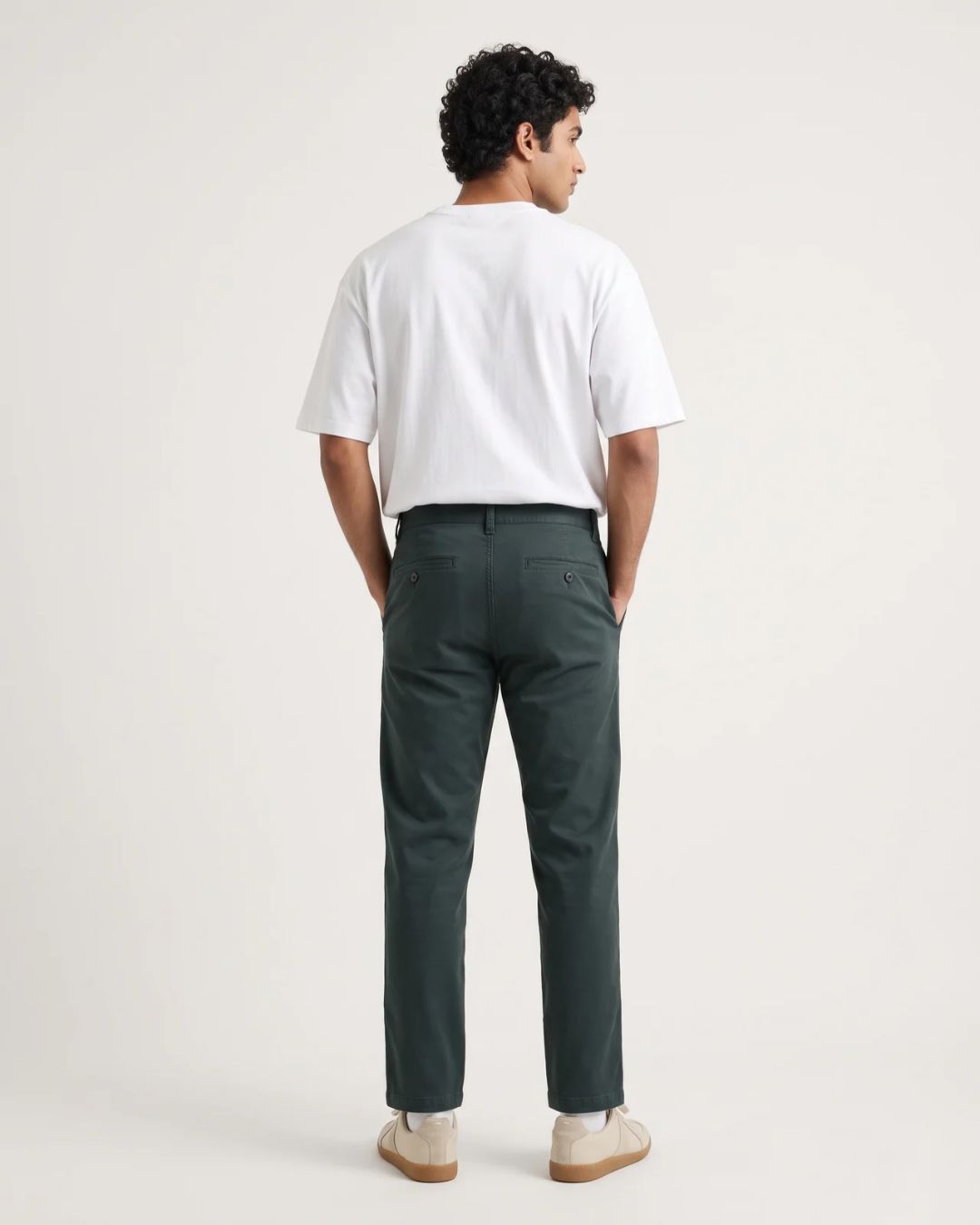 A man viewed from behind wearing a white t-shirt, dark green trousers, and beige shoes, standing against a plain off-white studio background.
