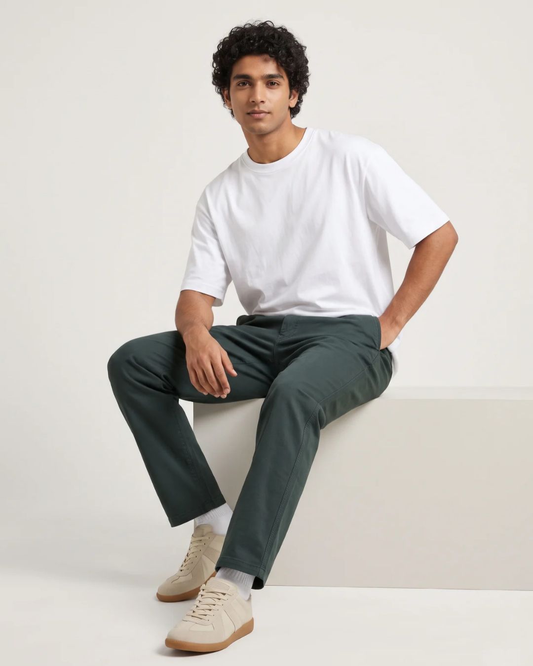 A man wearing a white crew-neck t-shirt, dark green trousers, and beige sneakers sits on a white platform against a light beige studio background.