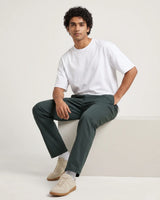 A man wearing a white crew-neck t-shirt, dark green trousers, and beige sneakers sits on a white platform against a light beige studio background.