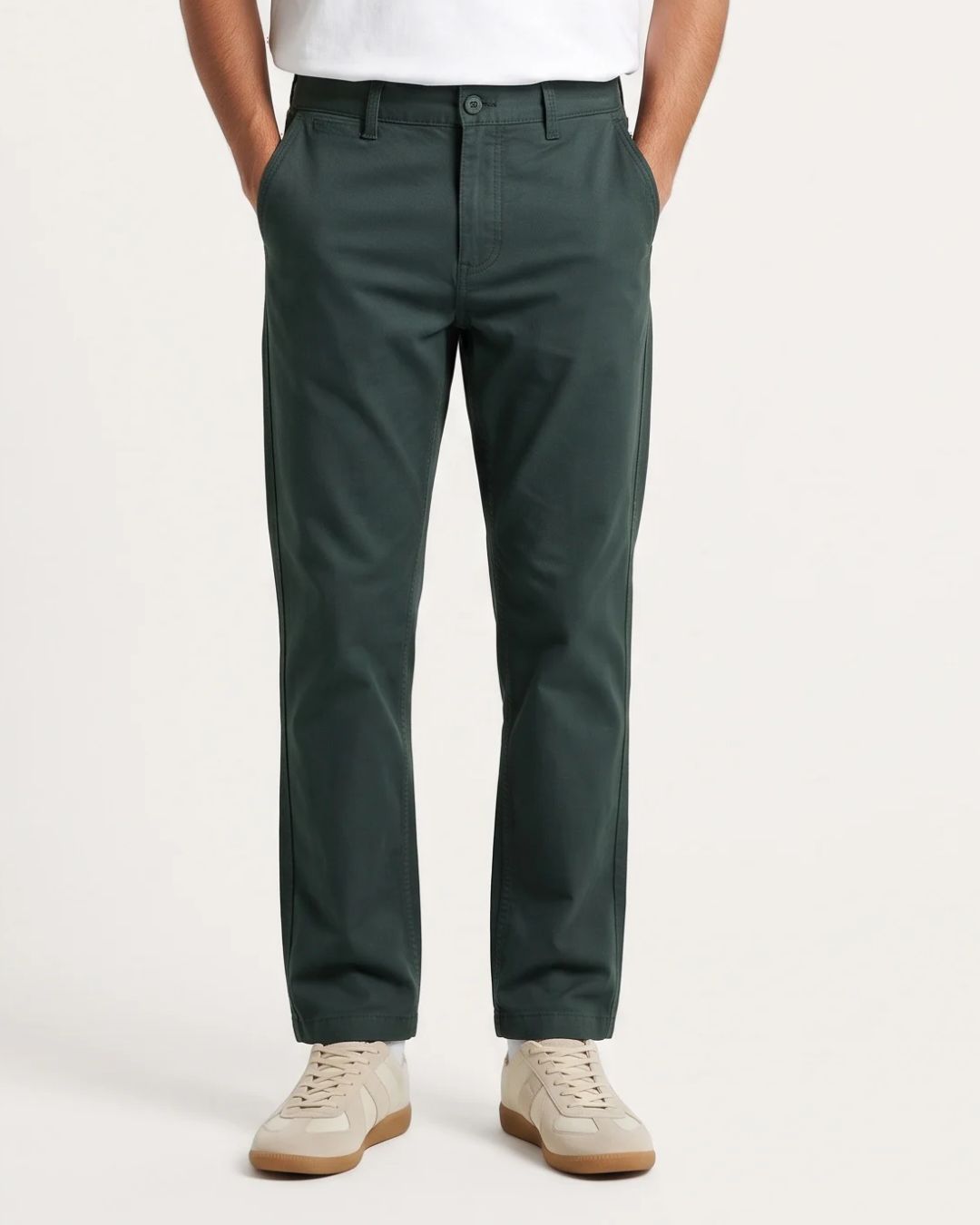 A person wearing dark green slim-fit chinos with a white shirt and beige sneakers, shown from the waist down against a light gray studio background.