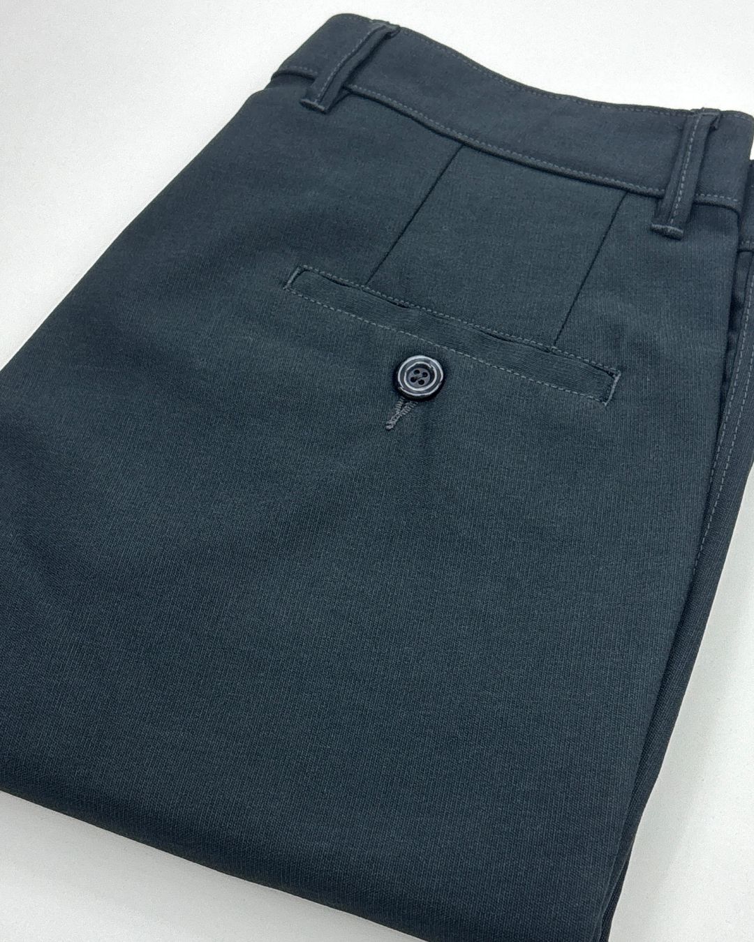 A close-up of folded dark charcoal gray trousers showing the waistband, belt loops, and a buttoned welt pocket.