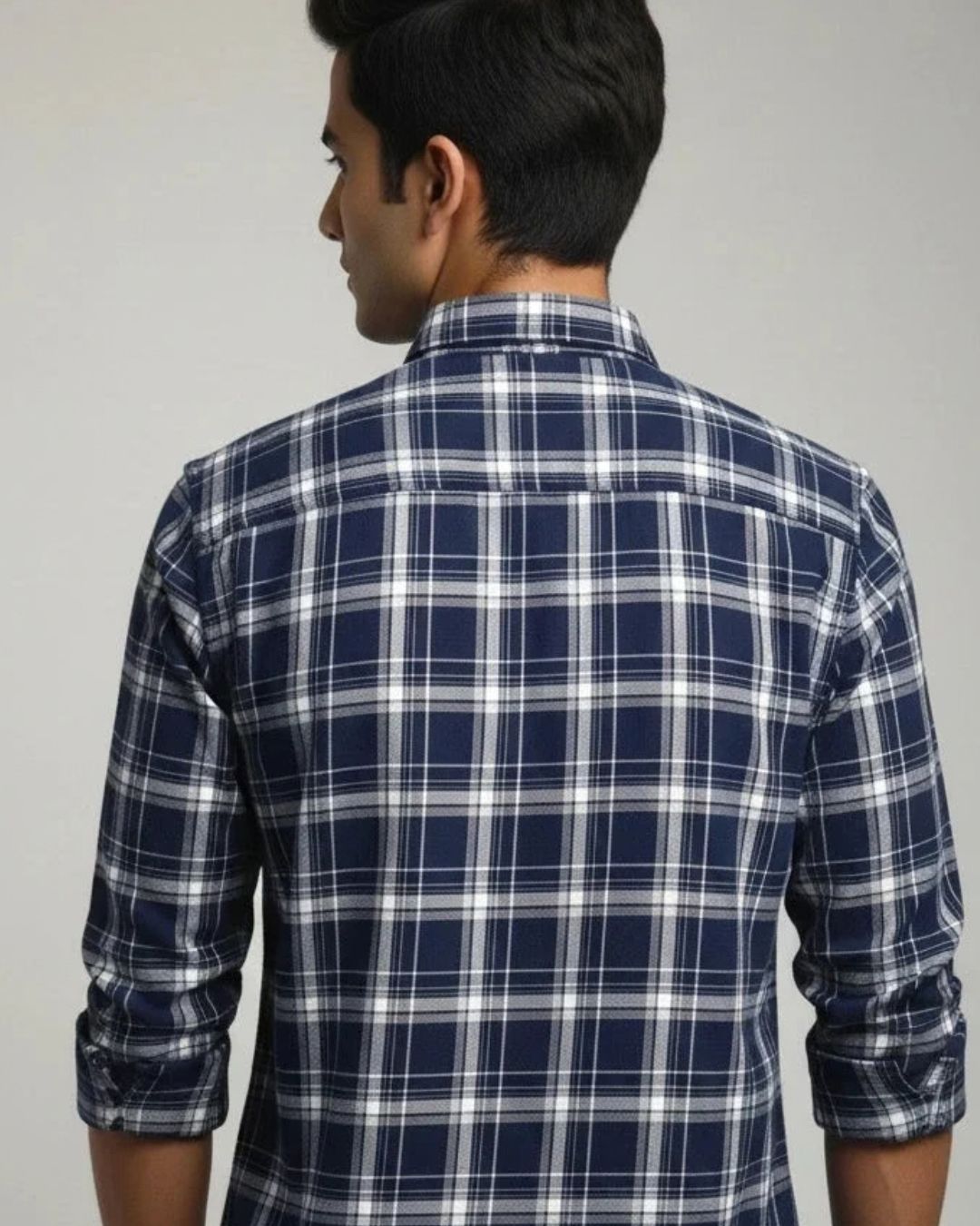 A man viewed from the back wearing a navy blue and white plaid long-sleeve shirt, with the sleeves rolled up, against a plain light gray background.