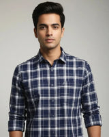 A man wearing a navy blue and white plaid long-sleeve button-up shirt, photographed against a neutral light gray background.