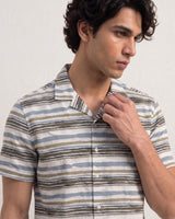 A man wearing a short-sleeve button-up shirt with horizontal stripes in beige, brown, blue, and white, posing against a light textured wall.