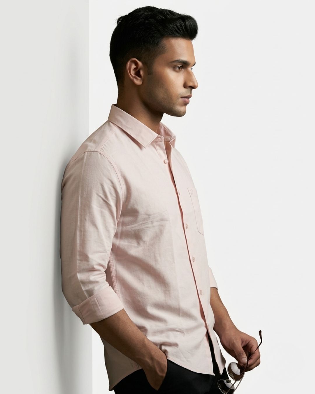 A man in profile wearing a light pink long-sleeve button-down shirt, leaning against a white wall and holding a pair of sunglasses.