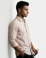 A man in profile wearing a light pink long-sleeve button-down shirt, leaning against a white wall and holding a pair of sunglasses.