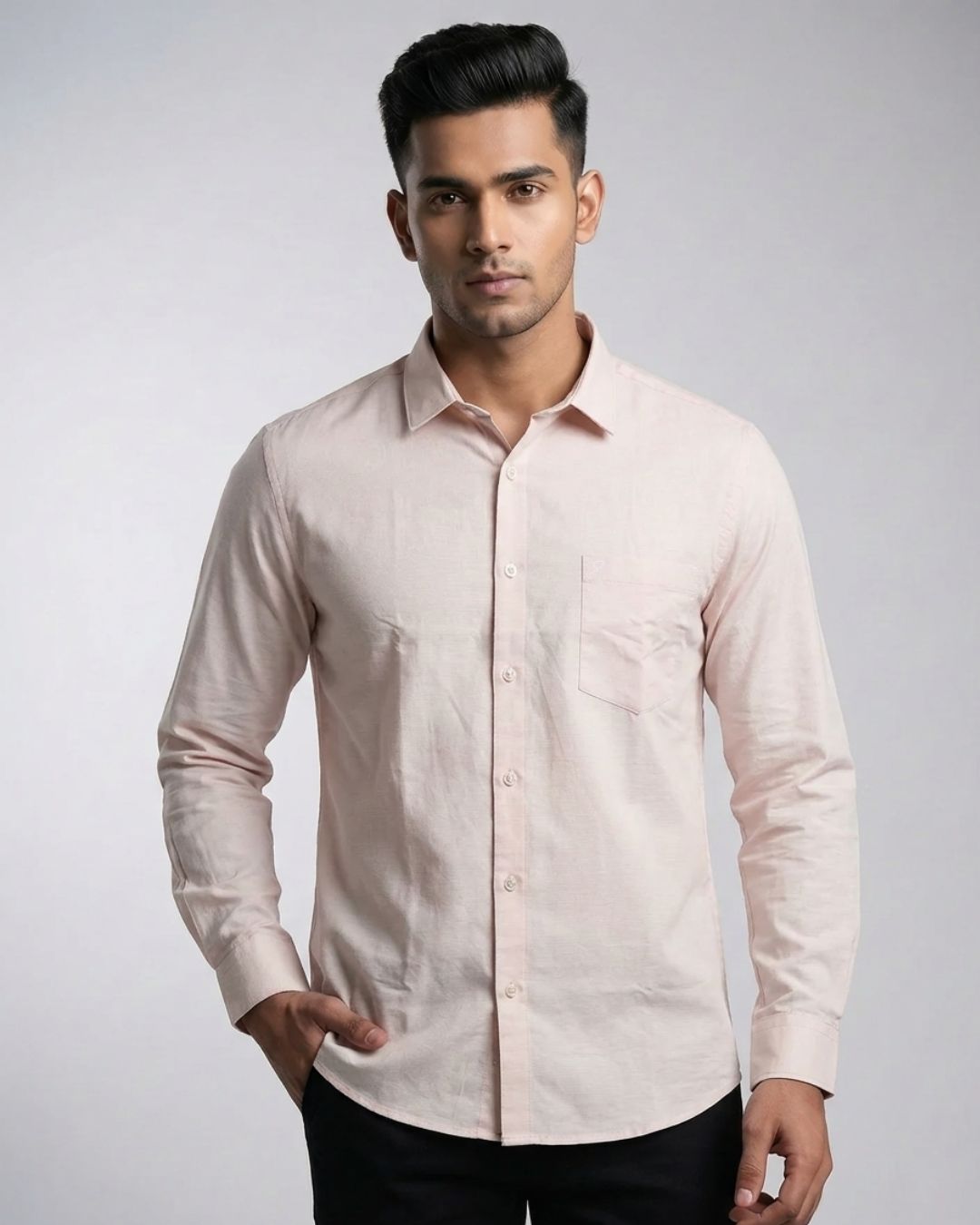 A man wearing a light pink long-sleeve linen shirt with a single chest pocket, paired with black trousers, standing against a light gray studio background.