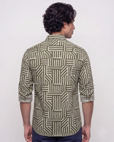 A man viewed from the back wearing a long-sleeve button-up shirt with an olive green and beige geometric block pattern, paired with navy blue pants, against a plain white background.