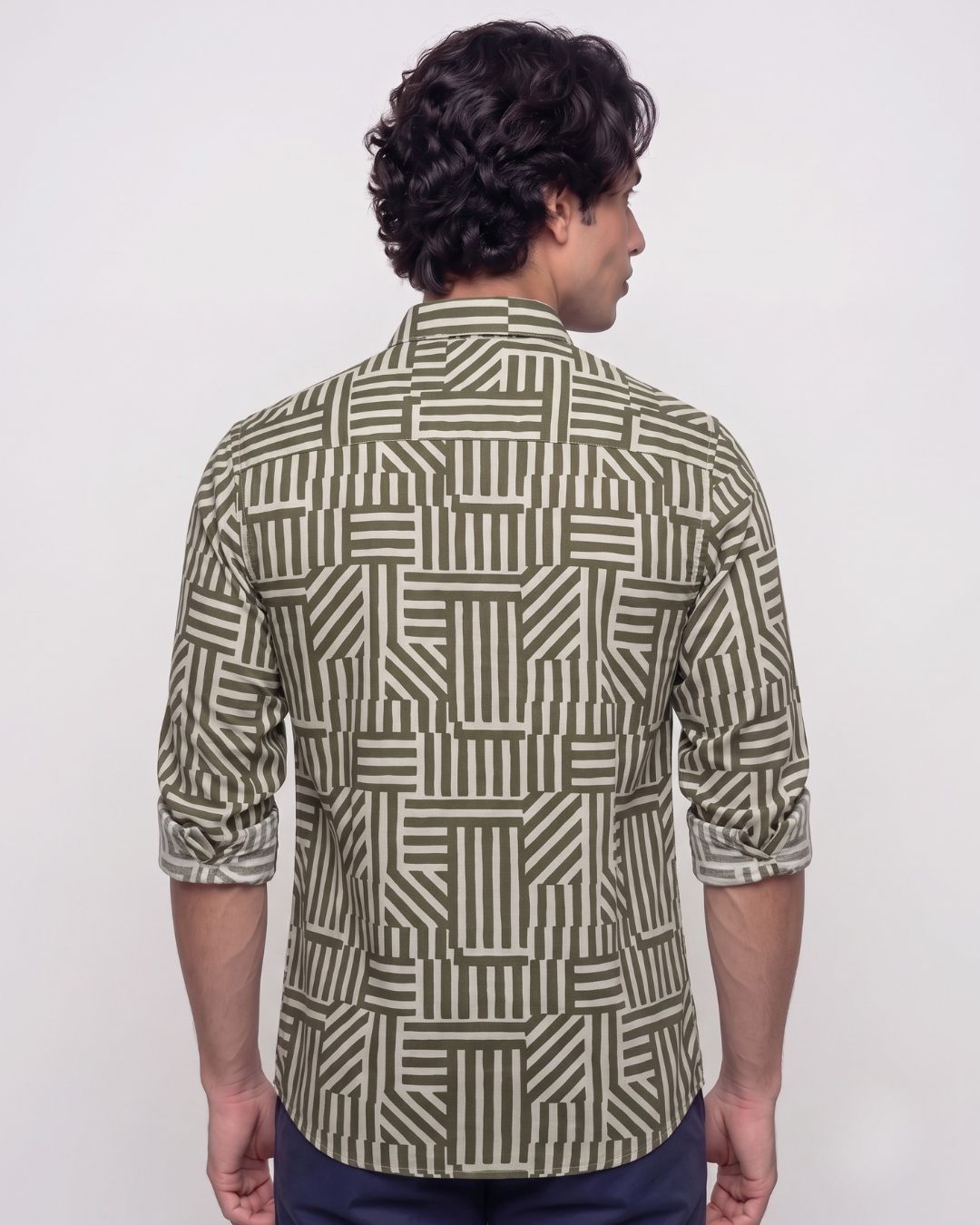 A man viewed from the back wearing a long-sleeve button-up shirt with an olive green and beige geometric block pattern, paired with navy blue pants, against a plain white background.