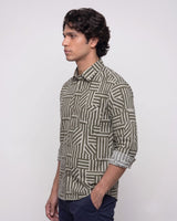 A man wearing a long-sleeve button-up shirt with a geometric striped pattern in olive green and beige, paired with navy blue trousers, shown in a side-profile view against a plain light gray background.