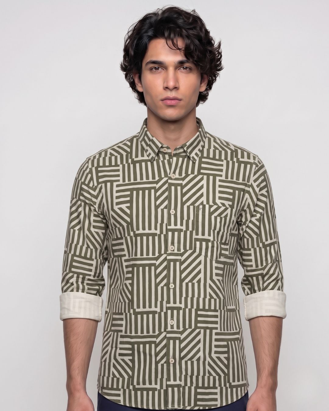 A man wearing a long-sleeve button-up shirt with a geometric striped pattern in olive green and beige, standing against a light gray background.
