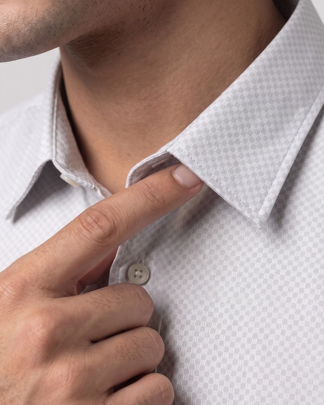Close-up of a man wearing a light gray and white micro-patterned dress shirt, adjusting the collar with his finger.