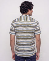 A man viewed from the back wearing a short-sleeve button-up shirt with horizontal wavy stripes in beige, blue, and brown tones, paired with navy pants, against a plain light gray background.