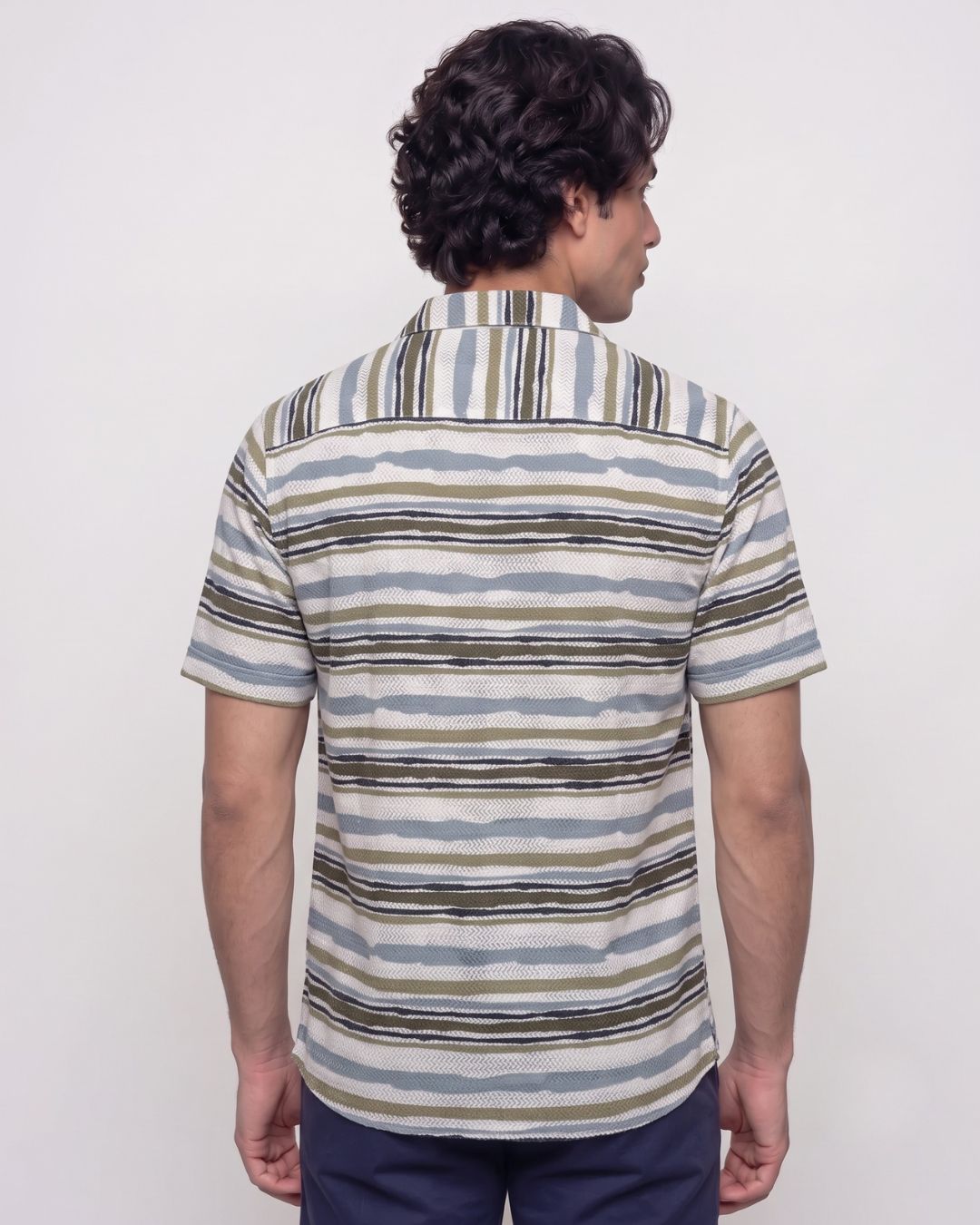 A man viewed from the back wearing a short-sleeve button-up shirt with horizontal wavy stripes in beige, blue, and brown tones, paired with navy pants, against a plain light gray background.