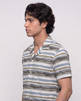 A man in profile wearing a short-sleeve button-up shirt with horizontal stripes in beige, brown, and light blue on a light gray background.