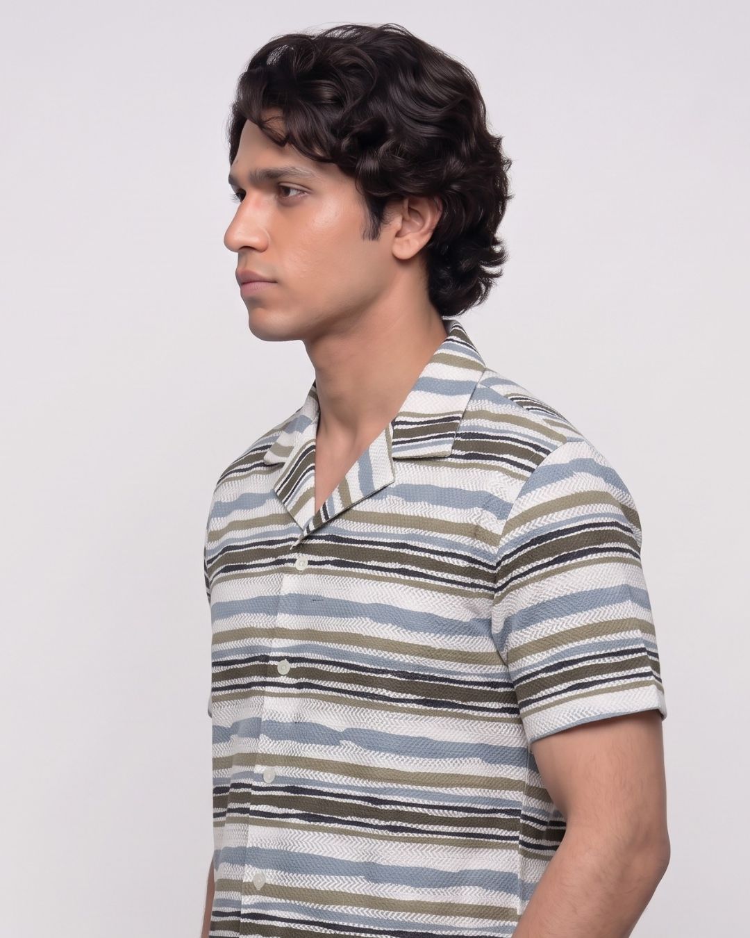 A man in profile wearing a short-sleeve button-up shirt with horizontal stripes in beige, brown, and light blue on a light gray background.
