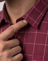 Close-up of a person wearing a burgundy plaid button-down shirt, pointing to the collar and button area.