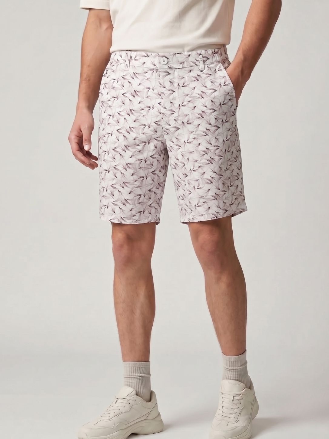 A person wearing white shorts with a delicate pink and gray leaf pattern, styled with a cream t-shirt, gray socks, and white sneakers against a light gray studio background.