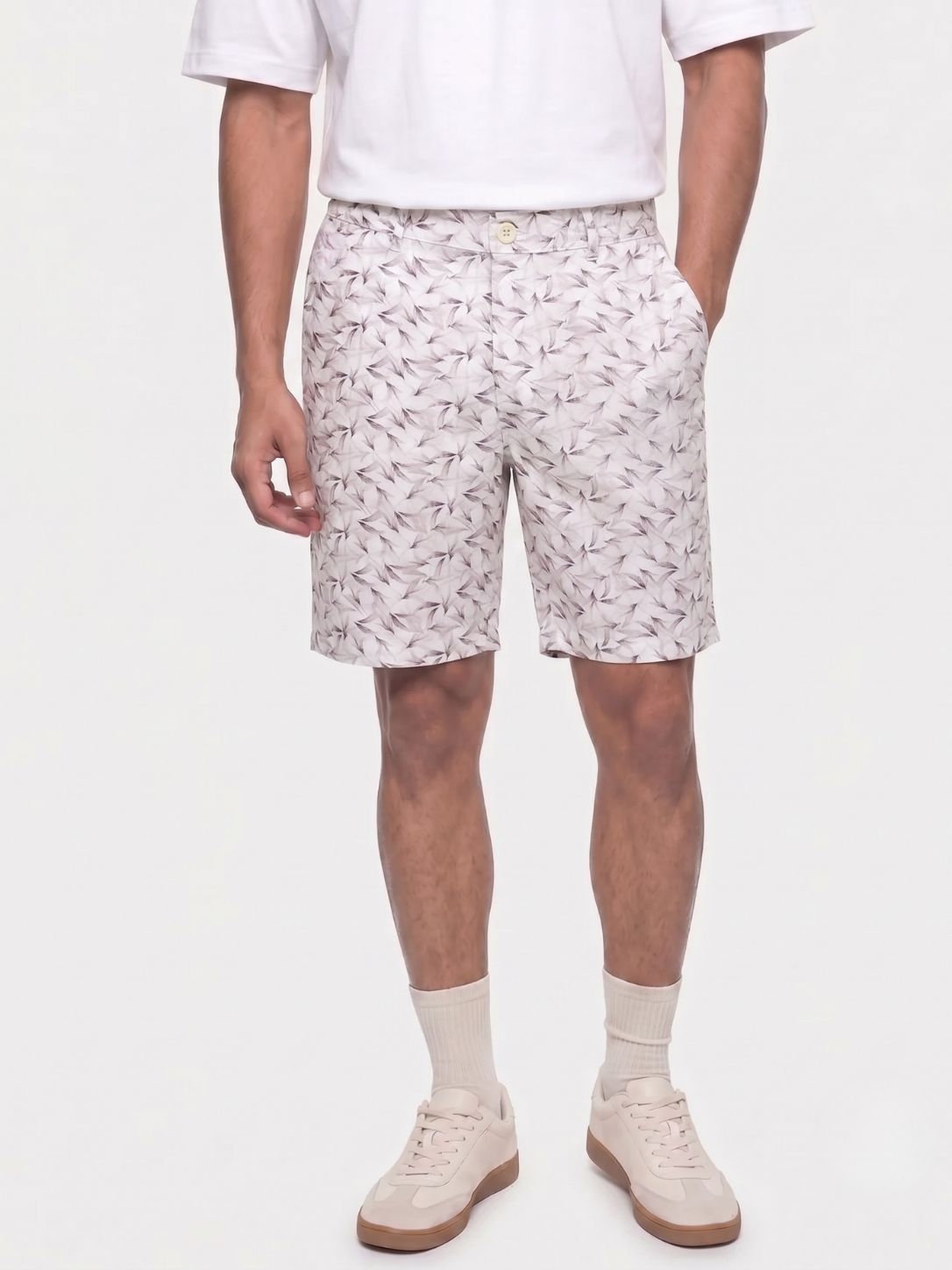 A person wearing light beige shorts with a small repeating dark leaf or feather-like print, paired with a white short-sleeve top, cream socks, and beige sneakers, photographed against a plain light background.
